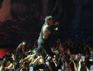 Performer with black clothing sings into a microphone while standing on a crowded audience's hands. Many attendees hold up phones.